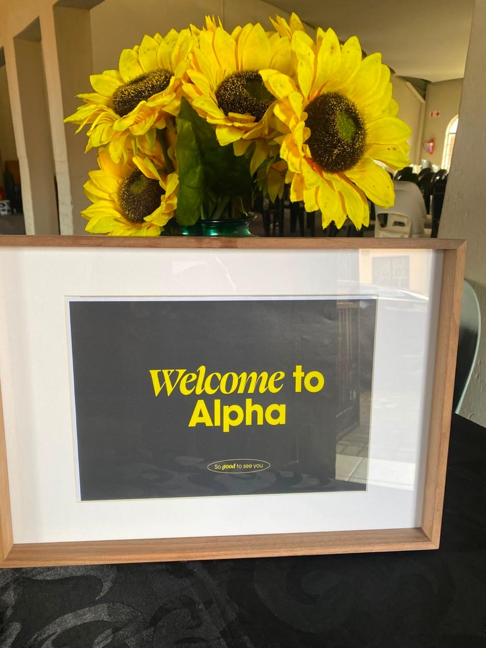 A framed “Welcome to Alpha” sign with sunflowers